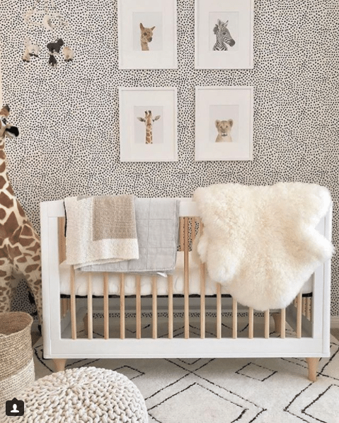 a nursery with animals themes. The crib has a white frame with maple wood dowels. hanging over the crib is a white fur blanket and two light colored towels. On the wall behind the crib is Leo's Spots wallpaper designed by Jillian Harris and manufactured by Milton & King. On the wallpapered wall are four framed pictures of a llama, zebra, giraffe and lion. To the left of the crib is a large stuffed giraffe toy. The floor has a large off-white soft rug with diamond shaped patterns in black outlines. Towards the front of the photo to the bottom left is a crocheted stool and wicker basket