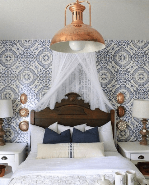 bedroom with blue Heritage Tiles Wallpaper by Milton & King on the wall behind a queen sized bed, two white end tables, each with a small lamp and a big bronze ceiling lamp 