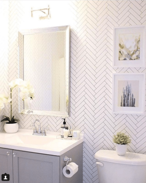 a bright white bathroom. The sink has gray wooden doors with a white top. A mirror with silver trim sits above the sink. On the wall is Tile Progress Wallpaper by Milton & King with two framed pictures of cactus.