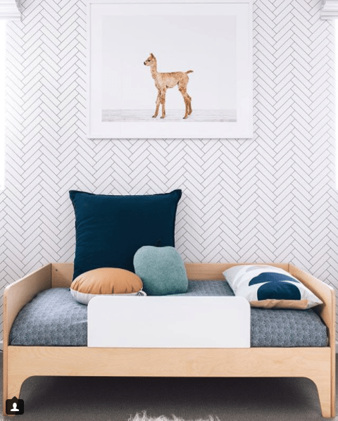 toddlers bedroom with Tile Progress wallpaper on the wall with a framed picture of a baby deer. Bed bed has a light wood bedframe and blue pattern bedding