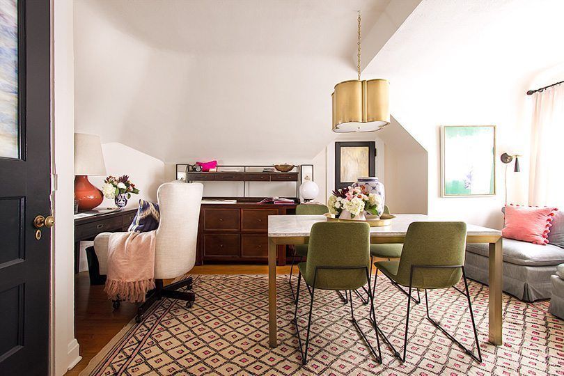 a wide photo of Nicole Balch's home office. On the left is her computer desk with a white upholstered chair. In the center of the room is a table with a floral arrangement on top with 4 olive green chairs. On the let is a gray sofa with pink pillows