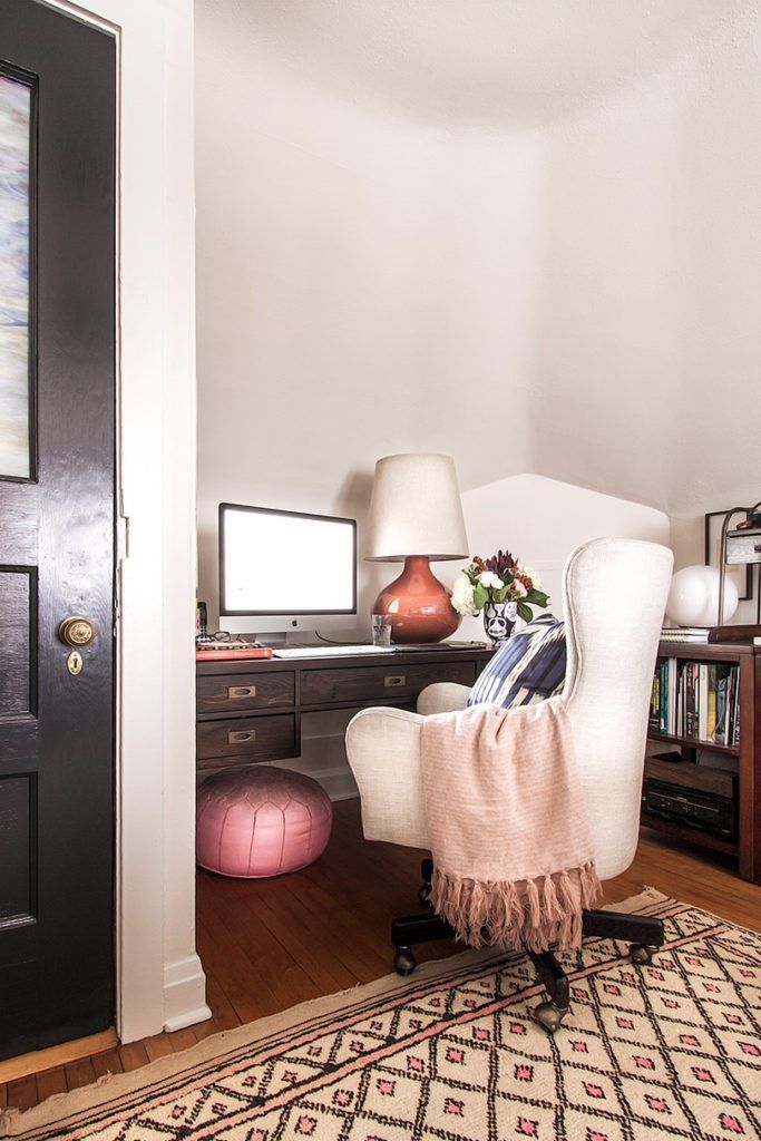 photo depicts a wide shot of an office desk with an orange lamp on the right and a computer monitor in the middle. In front of the desk is a white chair with a light colored blanker over the left arm. On the right is a small book shelf. On the floor is a tan colored carpet with small pink diamond shapes