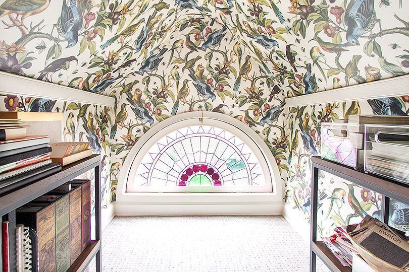 pictured is the closet with Orinthology wallpaper by Milton & King featuring birds of various shades of green, blue, yellow and brown among leafy branches. In the center is a half crescent stained glass window with two walnut bookshelves on the right and left side