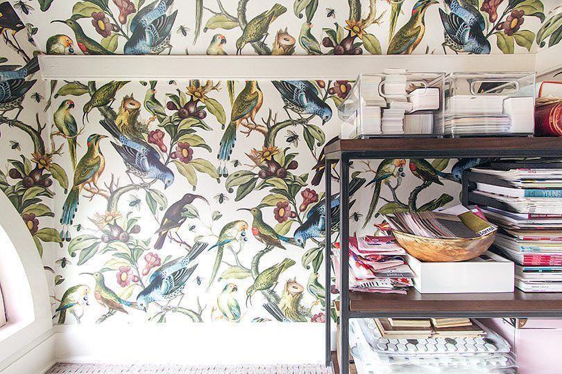 close up of Orinthology wallpaper by Milton & King with numerous colorful birds. On the right is a walnut shelf stacked with fabric samples, books and magazines