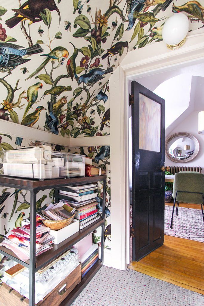 Pictured is a view out of the closet. On the left wall and ceiling is the Orinthology wallpaper by Milton & King. In front of the wall is walnut shelving with stacks of magazines, books and fabrics. There is a dark wood opened door leading into the office.