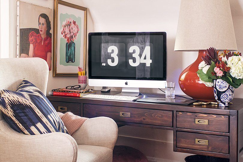 photo showing the time 3:34PM on the computer monitor that sits on a dark wooden desk. On the right of the desk is an orange lamp with a floral arrangement. To the left are two photos on the wall and in front of the desk is a white chair with a blue and white patterned pillow