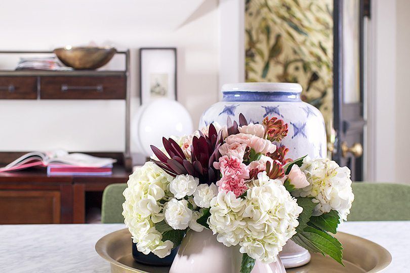 floral arrangement of white, pink and red flowers on top of marble topped dining table. In the background is a blurry image of an opened door with Orinthology wallpaper from Milton & King peeking through