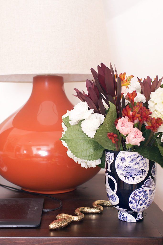 a close up shot of the orange ceramic lamp on the right side of the desk. A flat brass ornamental snake sits next to a black vase with blue print on it and a small arrangement of colorful flowers.