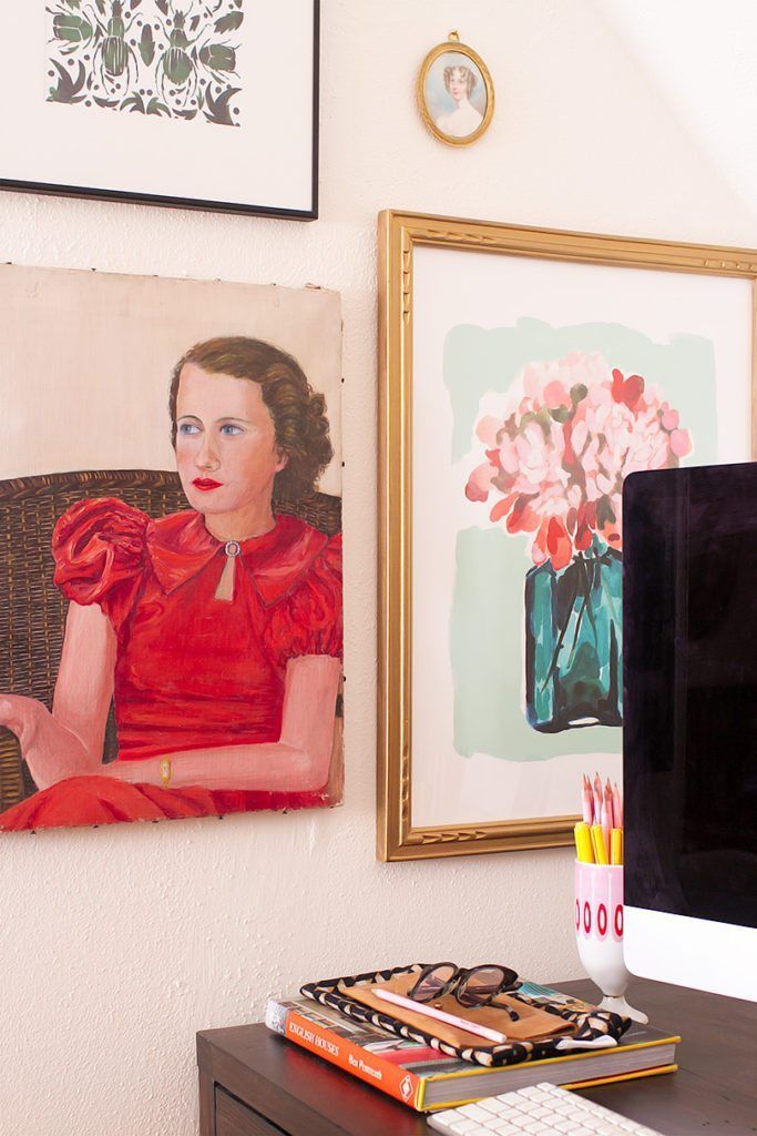 A close up shot of the art on the wall beside the desk. Show is a vintage painting of a woman from the 1930's. Above that is a print design of beetles. To the right of the beetle print is a small oval portrait of a woman. And below that is a gold framed painting of a pink floral bouquet in a blue vase.