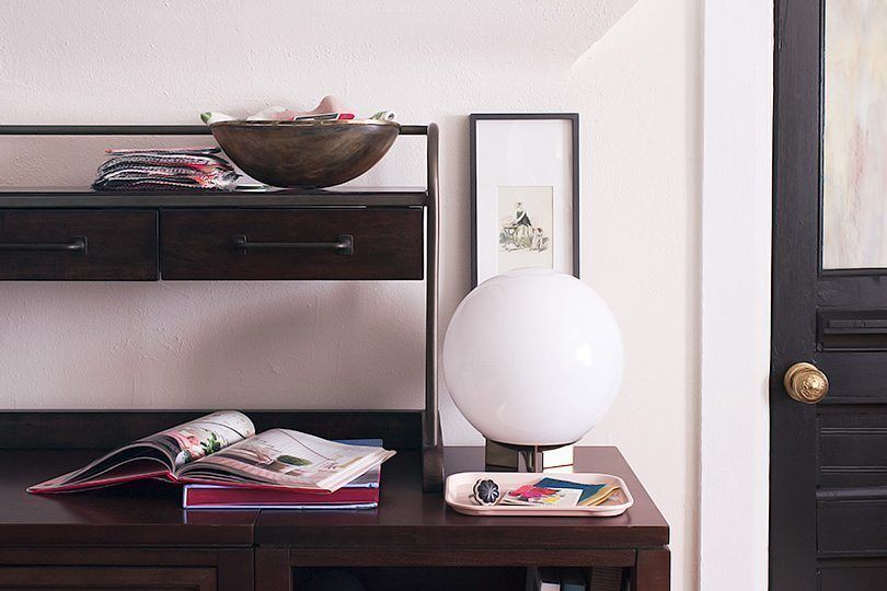 Close up of the right side of the desk. Also with a round light bulb lamp. on the top of the second tier of the shelf is a small pile of fabrics and wallpaper samples. On the lower part of the desk surface is an open book on top of a closed book.