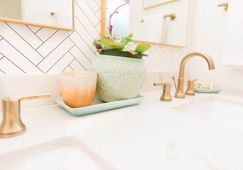 Close up shot of some plant and candle accents on the sink. On the right is brass plated faucet and handles and in the background is Tile Progress Wallpaper by Milton & King