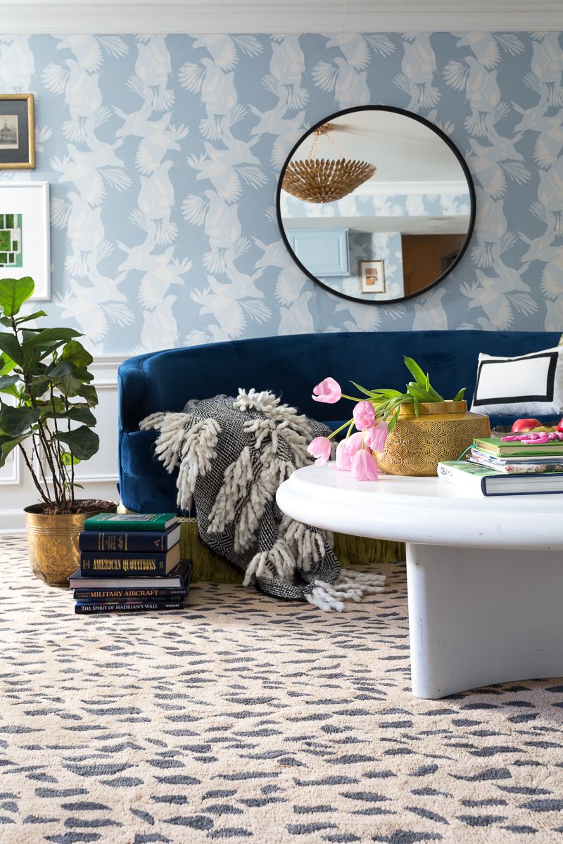 Jewel Marlow Basement Makeover • There is a leopard area rug a white round coffee table, a blue sofa and Magpie Wallaper from Mliton and King on the back wall with a big circular mirror mounted on the wall