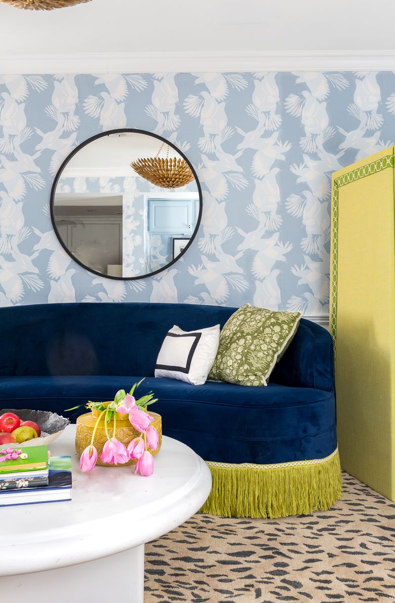 Jewel Marlow Basement Makeover • There is a leopard area rug a white round coffee table, a blue sofa and Magpie Wallaper from Mliton and King on the back wall with a big circular mirror mounted on the wall