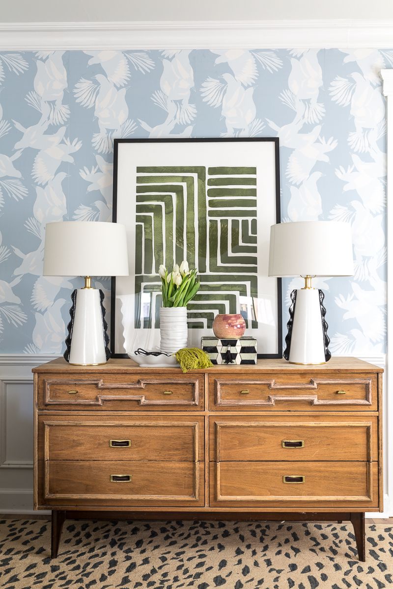 Jewel Marlowe Basement Makeover • a light brown wooden dresser has two white lamps on top and a piece of art and light blue magpie wallpaper from Milton & King