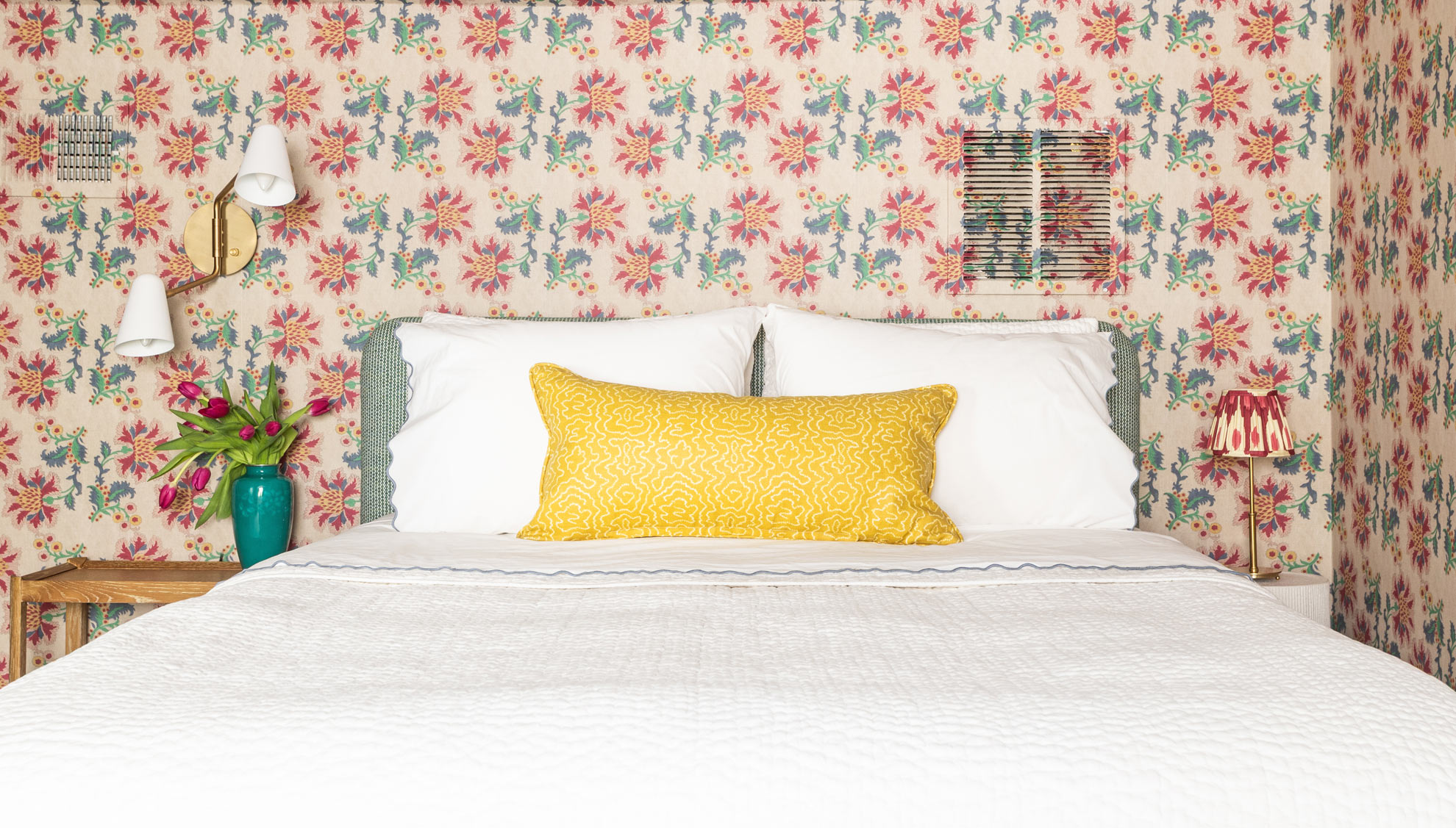 French Floral Wallpaper on the wall behind a bed with white bedding. A yellow pillow sits in the middle of the white pillows on the bed in this attic guest room.