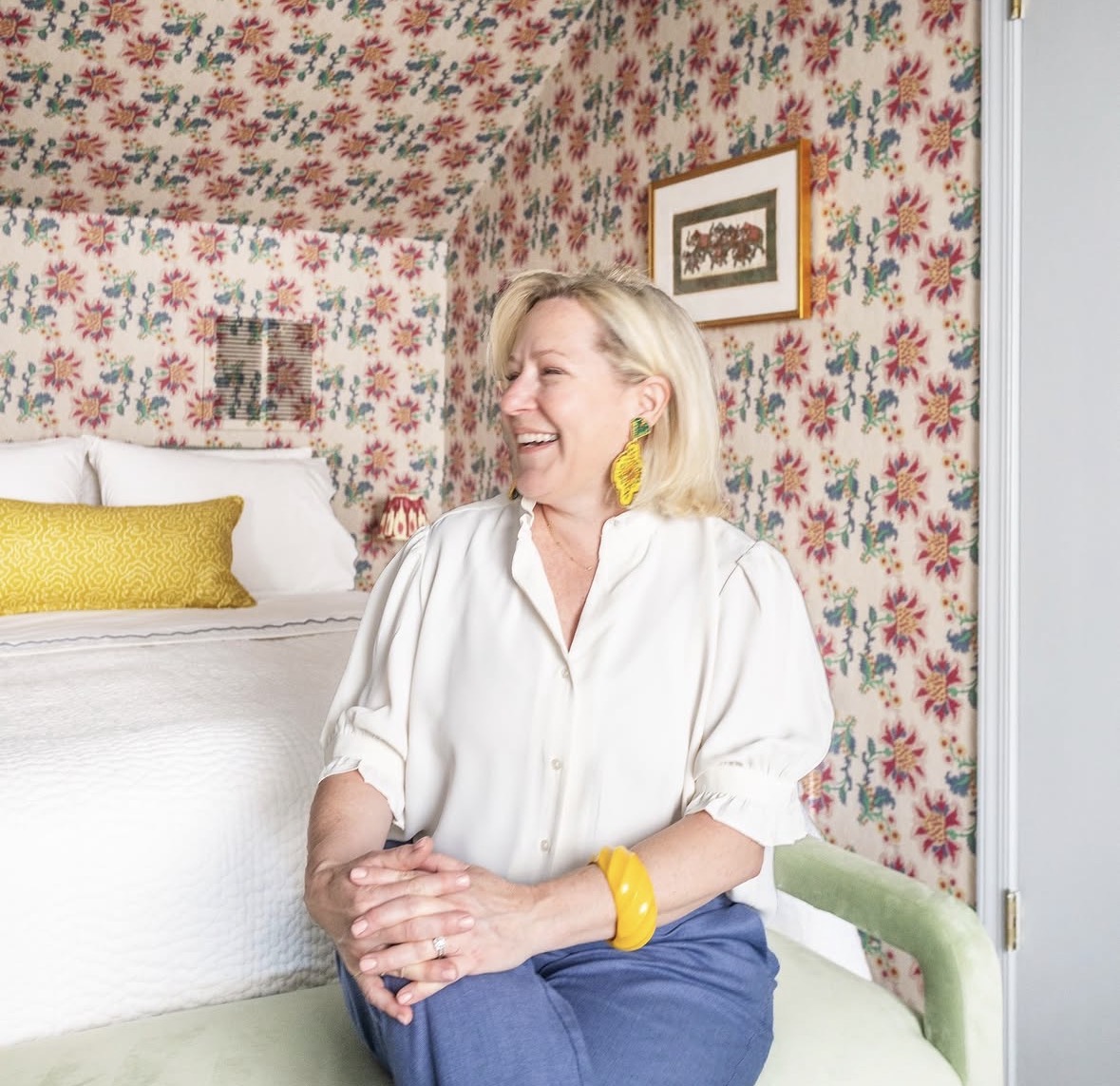 Kate Hougen from Mira Jean Interiors sitting on the light green bench at the foot a double bed with white sheets. In the background is a long yellow pillow on the middle of the bed and a colorful floral wallpaper.