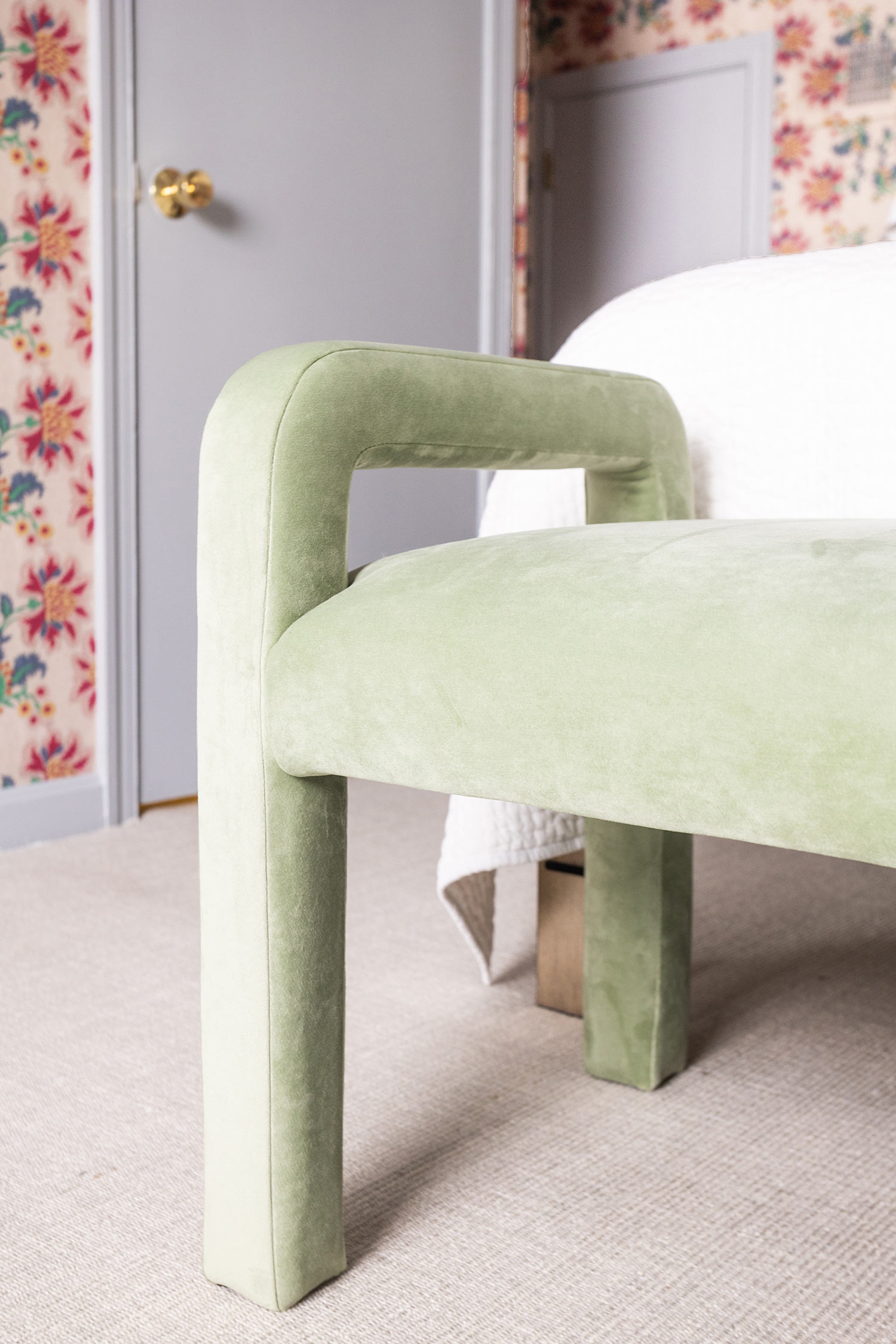light green velvet bench at the footer of the bed. in the background is a gray door with a gold handle and some colorful floral wallpaper